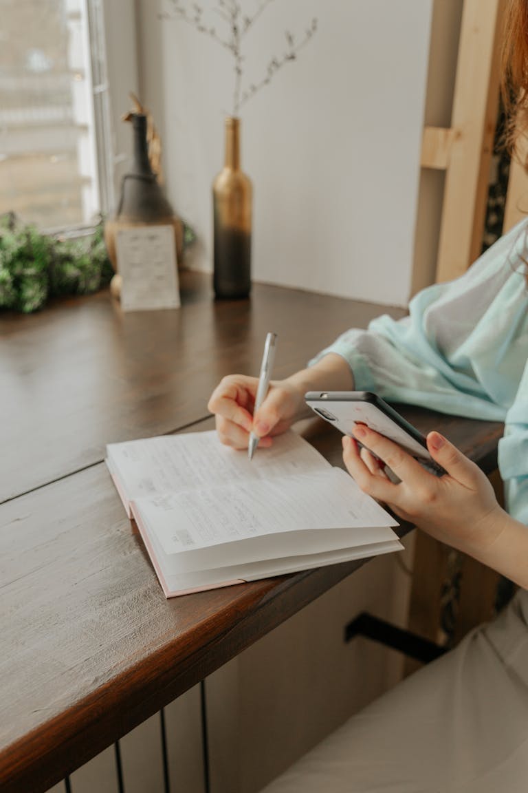anonymous female with cellphone and pen taking notes in diary at table in house