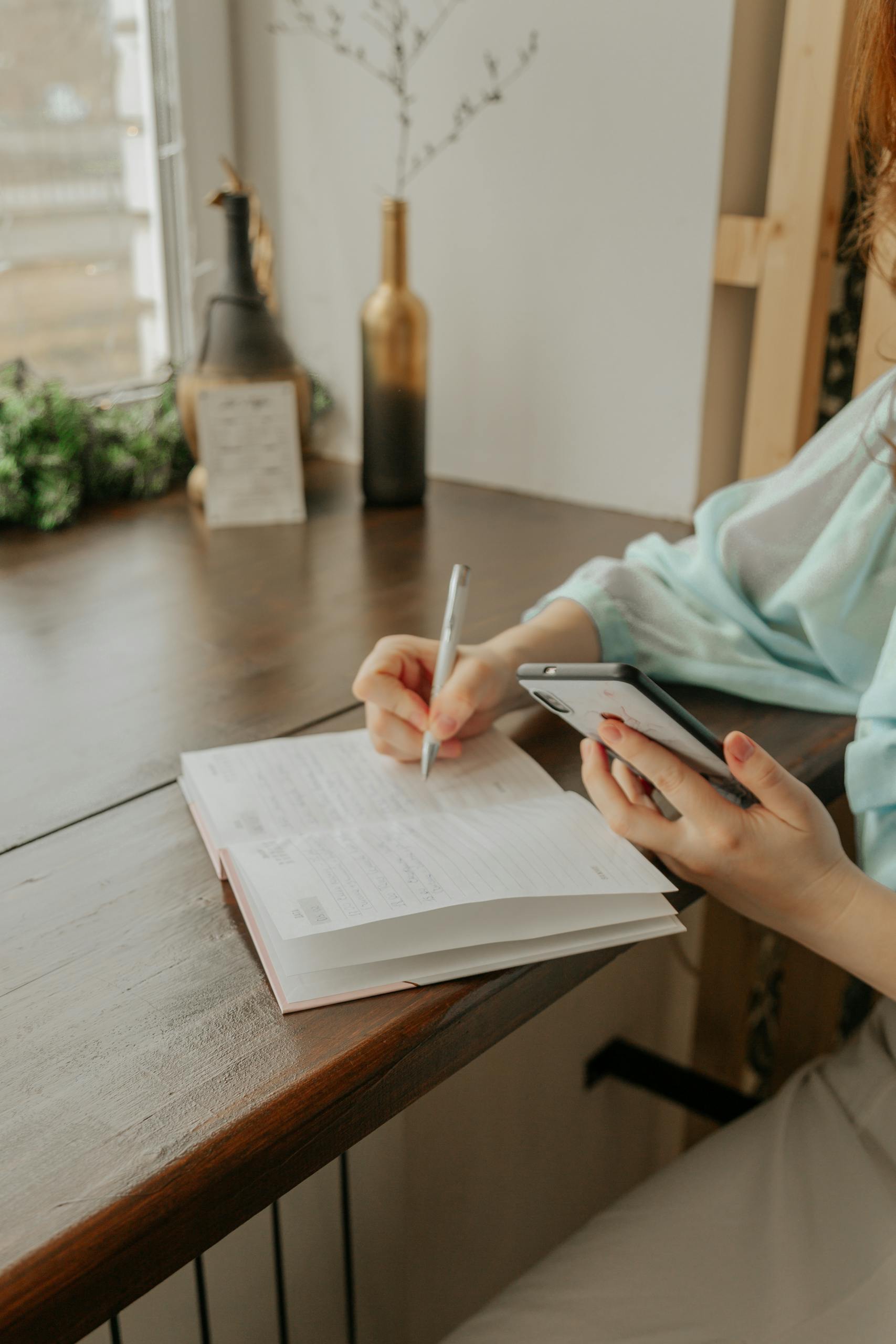 anonymous female with cellphone and pen taking notes in diary at table in house